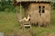 A naked woman sitting on a wooden chair in front of a hut.