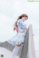 A woman in a blue and pink dress leaning against a fence.