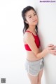 A woman in a red top and gray shorts leaning against a wall.