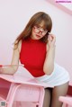 A woman in a red top and white skirt sitting at a pink desk.