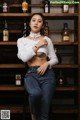 A woman sitting on a stool in front of a shelf of liquor bottles.