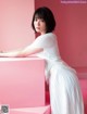 A woman in a white dress leaning against a pink wall.